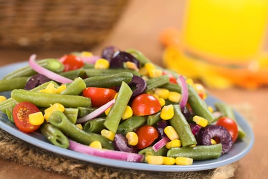 Fresh colorful vegetarian salad made of green beans, cherry tomatoes, sweet corn, black olives and red onions on blue plate with orange juice and basket in the back (Selective Focus, Focus one third into the salad) 