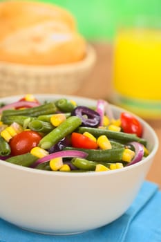 Fresh colorful vegetarian salad made of green beans, cherry tomatoes, sweet corn, black olives and red onions in bowl with orange juice and buns in basket in the back (Selective Focus, Focus one third into the salad) 
