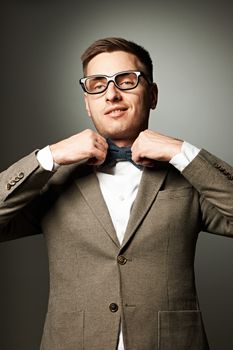 Confident nerd in eyeglasses adjusting his bow-tie against grey background