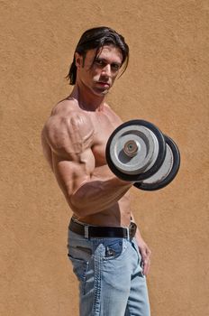 Muscular young bodybuilder shirtless outdoors in jeans, exercising bicep with dumbbell
