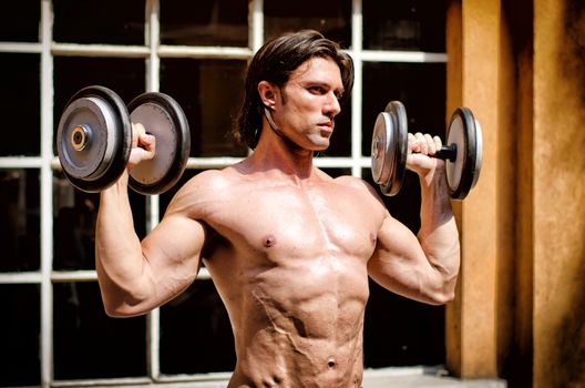 Muscular young bodybuilder shirtless outdoors, exercising biceps with dumbbells