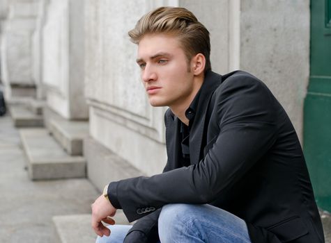 Attractive blond young man in jeans and jacket, sitting outdoors looking off camera