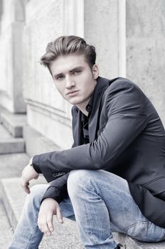 Attractive blond young man in jeans and jacket, sitting outdoors looking in camera
