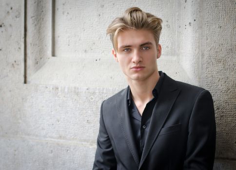 Attractive blond young man in shirt and jacket, standing against wall