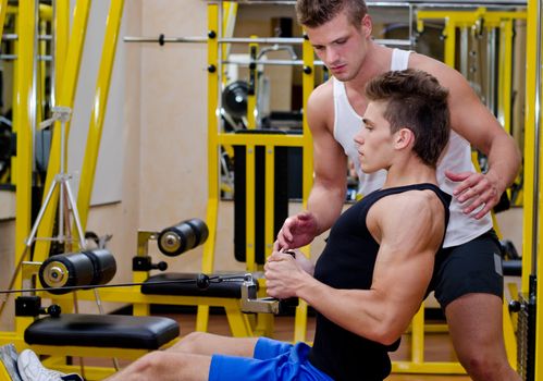 Personal trainer helping young male client in gym during workout on equipment