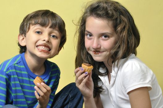 Two young children being silly putting chocolate frosting on their lips while eating cupcakes