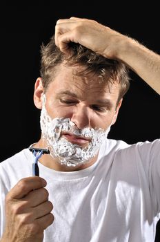 Man with shaving cream on face looks disgruntledly at his razor and scratches his head