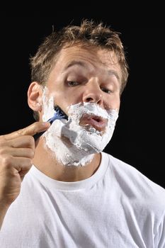 Closeup of Caucasian man with shaving cream on face making silly expression as he shaves his cheek on black background