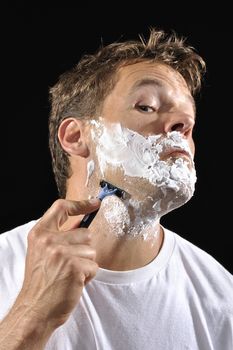 Handsome Caucasian man with shaving cream contorts face as he shaves his neck with black background