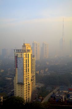 Vertical color capture of Bombay skyline, view obscured by the city haze and smog in the early hours of the day as the sun rises casting a golden hue over the tall buildings