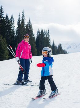 Happy little boy studying to ski in ski goggles and a helmet with his mother