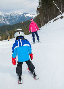 Happy little boy studying to ski in ski goggles and a helmet with his mother