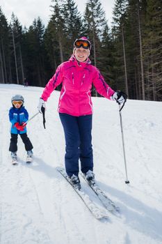 Happy little boy studying to ski in ski goggles and a helmet with his mother