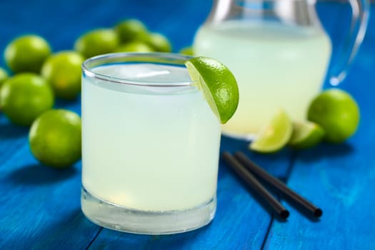 Freshly prepared refreshing lemonade out of limes with ice cubes in glass garnished with a lime wedge; limes, drinking straws and a pitcher in the back (Selective Focus, Focus on the lime wedge on the rim)