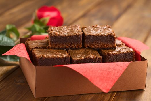 Freshly baked brownies in a brown paper box with red napkin, with red rose in the back (Selective Focus, Focus on the upper left brownie)