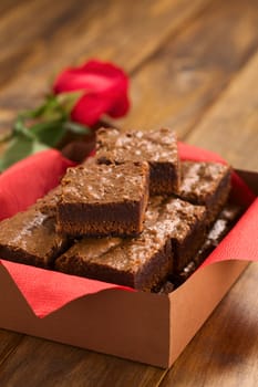 Freshly baked brownies in a brown paper box with red napkin, with red rose in the back (Selective Focus, Focus on the first brownie on the top)