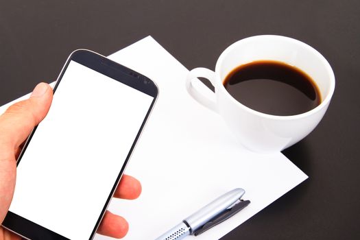 Hand showing smart phone with white, blank screen and paper, pen, coffee cup on wooden table.