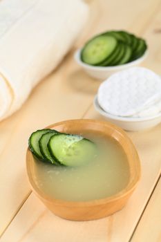 Homemade natural cucumber facial toner that hydrates, softens, soothes and cleanses the skin in a wooden bowl with cucumber slices, with facial pads and towel (Selective Focus, Focus on the front of the cucumber slices in the toner)