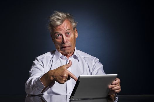 Emotional senior business man with an expressive face pointing to his laptop in horror and disbelief as he sits at a table against a dark studio background with copyspace