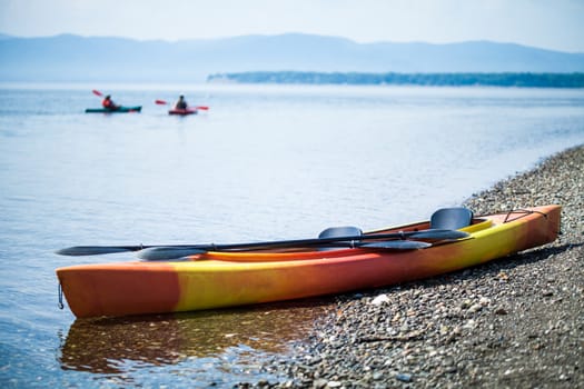 Orange and Yellow Kayak on the Sea Shore During a beautiful Day of Summer with Unrecognizable People Kayaking in the Background