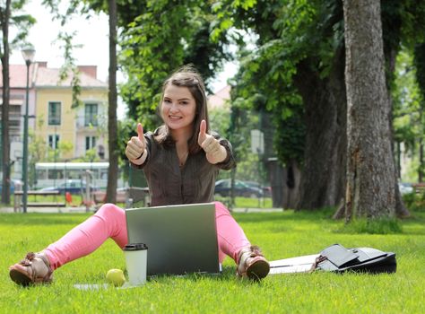 Happy young woman with a laptop rising her thumbs up, outside in an urban park.