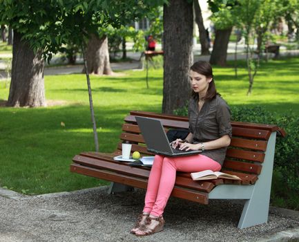 Young woman sitting on a bench in a park and working on a laptop.