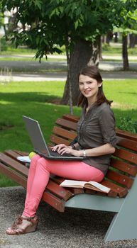 Young woman with a laptop sitting on a bench in ap ark and smiling to the camera