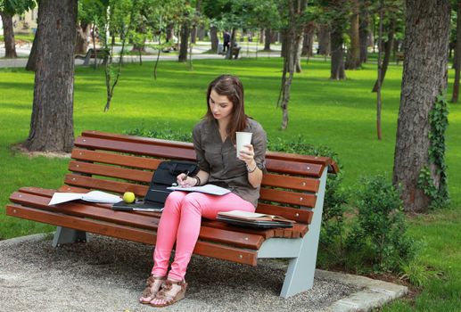 Young woman sitting on a bench in a park and writing something in a notebook while holding a disposable cup of coffee.