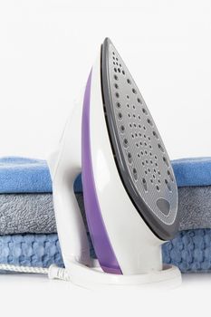 Standing steam iron and stacked blue bathroom towels, isolated on white background.