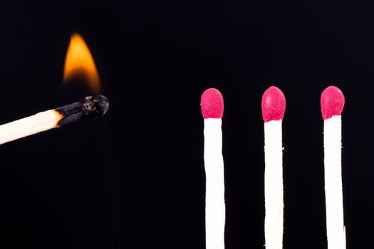 Burning match approaching red headed matches on dark background.