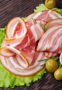 Arrangement of Smoked Ham Slices with Green Olives on Lettuce Leaves closeup on Dark Wooden background