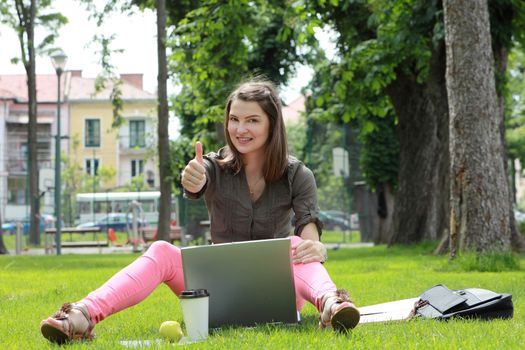 Happy young woman with a laptop rising her right thumb up, outside in an urban park.
