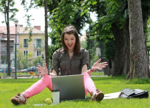 Happy young woman in front of a latop siting on grass outside in an urban park