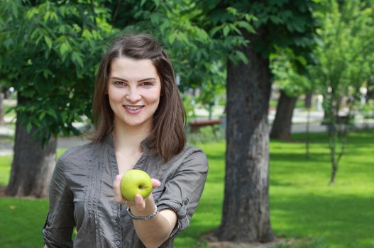 Young smiling woman offering a green apple outside in a park in summer.