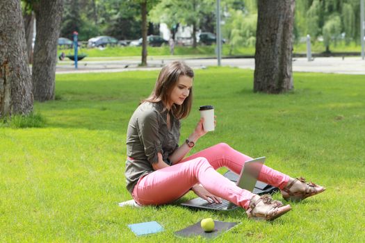 Young student woman holding a disposable cup of coffee while working on a laptop outside in an urban park.