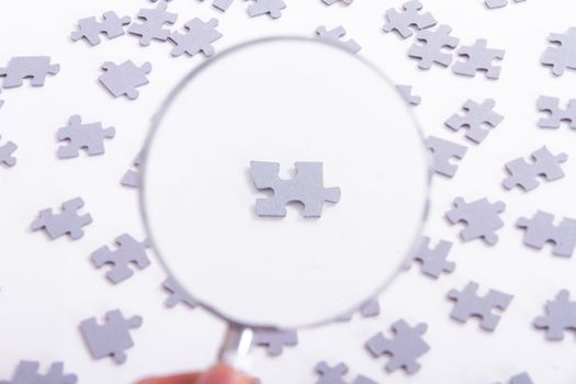 Hand holding classic style magnifying glass and examining single little puzzle piece among others on white.