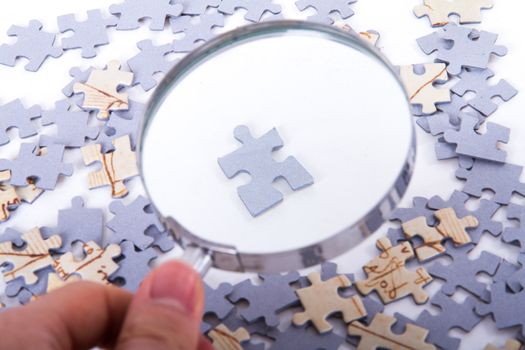 Hand holding classic style magnifying glass and examining single little puzzle piece among others on white.
