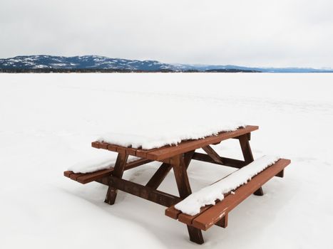 Closed for the season: beautiful lakeside campsite with wooden table and benches burried in snow and wide open flat of snowy frozen lake landscape with pristine white snow on a cold winters day