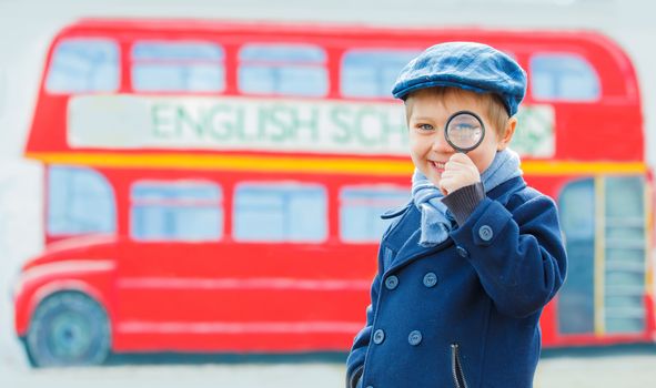 Little detective. Funny boy looking through magnifying glass near english schoolbus