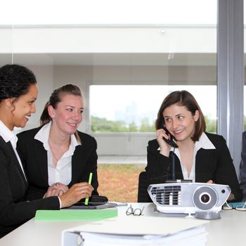 Three successful businesswomen or partners in a meeting sitting together at a table smiling in satisfaction as one talks on the telephone watched by the others