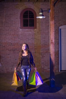 Excited Pretty Mixed Race Young Adult Woman Walking in the Evening Holding Shopping Bags with Brick Wall Background.