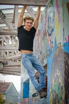 Attractive blue eyed, blond young man in jeans and black t-shirt hanging from concrete structure
