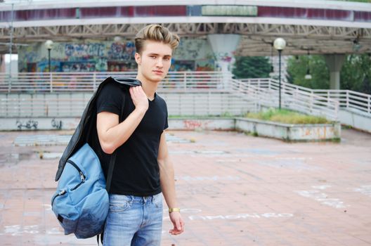 Handsome blond, blue eyed young man outdoors with ruck sack on shoulder, t-shirt and jeans