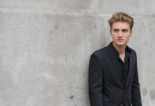 Attractive blue eyed, blond young man leaning against stone wall, looking to a side, large copy space