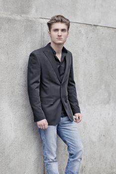 Handsome blond young man, blue eyes, leaning against white, rough stone wall, looking up to a side