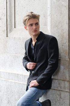 Attractive blue eyed, blond young man leaning with his back against white wall outdoors
