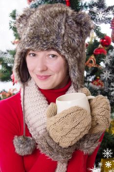 Smiling woman in fluffy hat and mitten under Christmas tree with cup