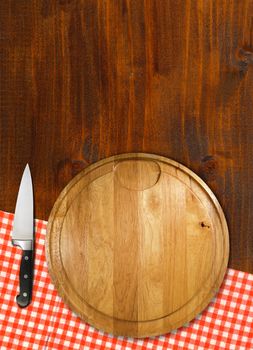 Round cutting board with red checked tablecloth and kitchen knife on brown wooden table