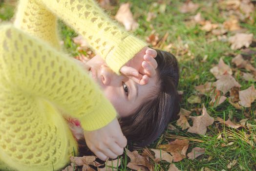 Young woman laying in grass with a bunch of fallen leafs, covering one eye