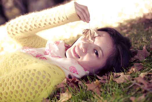 Young woman laying in grass with a bunch of fallen leafs, covering one eye with leaf